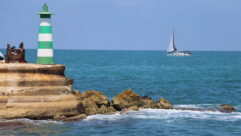 A green and white striped lighthouse is situated on a rocky outcrop in the sea. The lighthouse is built on a sturdy stone platform. Nearby, a sailboat with white sails glides across the vibrant blue waters, with the horizon stretching into a clear blue sky. The rocks around the lighthouse show signs of wear from the sea, with waves gently lapping at their base. The scene captures a serene maritime setting.